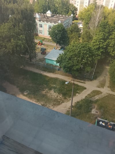 The image is an aerial view of a playground area next to a building, likely a kindergarten or school, in the city of Zhlobin, Belarus. The building has a light-colored brick facade and a grey roof with decorative turrets. Adjacent to the building is a playground with a small, colorful playhouse and other playground equipment. A fence separates the playground from a dirt path and grassy areas. A street lamp with a single light is visible in the lower right quadrant of the image. In the far background, several tall apartment buildings can be seen amidst lush green trees. A row of garbage bins is also present near the bottom right corner of the image, some of which contain visible trash. The overall scene depicts a typical urban environment in Belarus, with a focus on a recreational space for children.