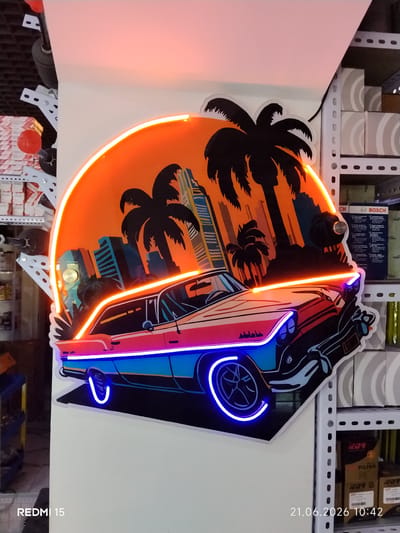 The image shows a neon sign shaped like a classic car with palm trees and a city skyline in the background, set against a bright orange sunset. The sign is mounted on a white wall in what appears to be a retail or storage space. Shelving units with boxes are visible to the right of the sign. The overall impression is a vibrant, retro-themed decorative piece. The image includes a timestamp and model name at the bottom: "REDMI 15 21.06.2026 10:42". There is no visible indication of the city of Yovon, Tajikistan.