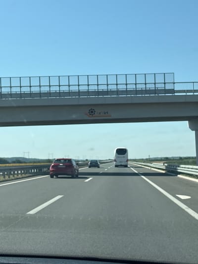 An eye-level, forward-facing shot captures a highway scene under a clear blue sky in Hrtkovci, Serbia. The foreground is dominated by the asphalt road, marked with white lines indicating lanes and a dashed line on the left. In the middle ground, a red Audi sedan is seen from the rear, traveling in the right lane. Further ahead, a white bus occupies the same lane, while a darker colored car is visible in the left lane. Overhead, a concrete overpass stretches across the highway, featuring a metal railing and a section of advertising or signage with a gear-like logo and text that appears to read "Azvirt". Beyond the highway, a flat landscape unfolds with fields and distant power lines, suggesting a rural or semi-rural setting. The overall impression is one of transit and travel on a sunny day.