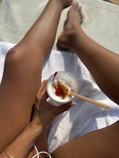 A person is holding a drink garnished with a dried apple and a cherry in their lap. They are sitting on a white towel over a cushioned lounge chair. The person's tan legs and feet are visible. The drink is white and frothy, with a cinnamon-colored powder sprinkled on top. A tan straw sticks out of the drink.
