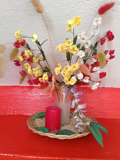 A floral arrangement sits on a wicker tray in front of a red wall and white wall. The arrangement includes several types of artificial flowers in red, yellow, and white, as well as some dried decorative grasses. A red candle is placed on the wicker tray, along with some straw-like material and green artificial leaves. The setting appears to be indoors, possibly a home or a shop. The image is a still life composition with no people present. The specific location in the city of Tebras, Latvia is not discernible from the image itself.