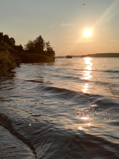 This image depicts a serene sunset over a body of water, likely a lake or a large river. The sun is a prominent feature, positioned in the upper right quadrant of the frame, casting a warm, golden glow across the scene. Its rays create a shimmering reflection on the rippling surface of the water, adding a sense of dynamism to the otherwise tranquil setting.
The shoreline on the left is characterized by lush vegetation, with trees and bushes creating a natural boundary. In the distance, the opposite shore is lined with trees, forming a dark silhouette against the bright sky. There are a few small boats or watercraft visible on the water, suggesting recreational activity, and a faint speck in the sky indicates an airplane, a subtle sign of human presence in the broader environment.
The time of day is clearly sunset, given the warm, low-angled light and the colors of the sky, which transitions from a pale blue at the top to soft oranges and yellows near the horizon. The weather appears to be calm, with gentle waves on the water. The overall mood evoked by the image is peaceful and contemplative, capturing the beauty of nature at the end of the day.
The location is identified as Boltyino, Mytishchinsky District, Russia, which provides geographical context. There is no visible text within the image.