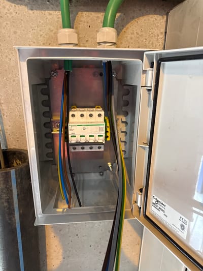 The image shows an open electrical distribution box mounted on a concrete wall in Kallo, Belgium. The box is white plastic and contains a Schneider Electric miniature circuit breaker labeled "C1". Several electrical wires, including green, yellow, blue, brown, and black, are visible entering the box through cable glands. One green cable appears to be conduit. The interior of the box has a ribbed mounting structure. The lid of the box, which is open, displays a label with "Schneider Electric NSYPLM32G" and a CE marking. The environment appears to be an outdoor or utility space, with rough concrete walls and some industrial-looking pipes or conduits visible in the lower left.