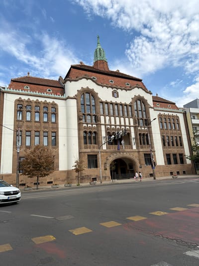 The image captures a view of the imposing Debrecen City Hall in Debrecen, Hungary, on a partly cloudy day. The building, characterized by its ornate Art Nouveau architecture, features a cream-colored facade with brown stone detailing around windows and arches. A prominent arched entrance is visible in the center, with two women walking on the sidewalk in front of it. The roof is tiled in red, and a green spire crowns the building. In the foreground, a street with tram tracks runs across the scene, and a white car is parked on the left side of the road. The sky is a vibrant blue with scattered white clouds.