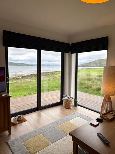 An indoor scene in Sconser, United Kingdom, shows a modern room with expansive windows offering a panoramic view of a natural landscape. The room features light-colored walls and ceiling, with black framed windows equipped with black blinds. The flooring is light wooden planks, partially covered by a rectangular rug with a geometric pattern in shades of grey, yellow, and cream.

To the right, a wooden table holds a black remote control, a small black device, and a red box with the word "UNO" visible on its side. Above the table, a fabric lampshade with a woven base hangs from the ceiling. Near the windows, a woven basket contains folded items, and a small potted plant sits on the windowsill. In the foreground at the bottom, a wooden console or table displays a small screen, some glasses, and decorative white ceramic items on a small brown mat.

Through the windows, a body of water (likely a loch or sea) is visible, with a rocky shoreline. Beyond the shore, lush green grassy land stretches towards distant, rolling hills or mountains under an overcast, cloudy sky. A wooden deck or patio area is visible directly outside the windows, suggesting an outdoor extension to the living space.

No people are present, and no specific activity is taking place, but the setup suggests a comfortable living area designed to appreciate the scenic surroundings. The daylight indicates it is daytime, although the cloudy weather gives a soft, diffused light. The overall atmosphere is tranquil and inviting, connecting the modern interior with the rugged, natural beauty of the Scottish landscape.