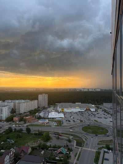 The image captures an aerial view of a city, likely Boldino, Russia, during a dramatic sunset with heavy clouds. The sky is a mix of dark, ominous storm clouds and bright, golden sunlight breaking through on the horizon. In the foreground, the reflective facade of a modern building dominates the right side of the frame, mirroring the sky. Below, a dense forest separates the city from the distant horizon, where a cityscape of apartment buildings and industrial structures can be seen. Closer to the viewer, a large retail complex with a parking lot filled with cars is visible, surrounded by residential buildings. A multi-lane road with a roundabout is also present, with several cars navigating the intersection. The overall scene suggests a blend of urban development and natural landscape, with the weather creating a striking visual contrast.