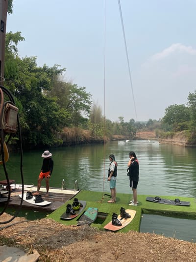 The image depicts a wakeboarding park located in Tambon Nam Phrae, Thailand. The scene shows three individuals on a dock area near a body of water, presumably a lake or pond used for wakeboarding. The individuals are wearing casual clothing and life vests, suggesting they are preparing for or have just finished wakeboarding. Wakeboards are also visible on the dock. The background consists of lush green vegetation and trees lining the banks of the water, typical of the natural environment in this region of Thailand. The sky is slightly hazy, indicating possible atmospheric conditions in the area. Overall, the scene conveys a recreational activity taking place in a natural setting in Thailand.