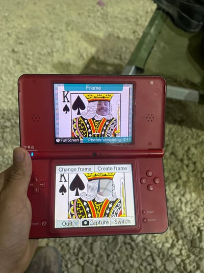 A person is holding a Nintendo DS handheld video game console in their left hand. The console's top screen displays a "Frame" of the King of Spades playing card, with a person's face superimposed onto the card's king. The bottom screen displays another "Frame" of the King of Spades playing card, but the person's face is not superimposed. The console has options for "Change frame," "Create frame," "Quit," "Capture," and "Switch." The console also has buttons for "Start" and "Select." The person is holding the console in a rough outdoor setting, with the ground beneath them being a rough, dry, pebbled surface. The location is in Tze'elim, Israel.