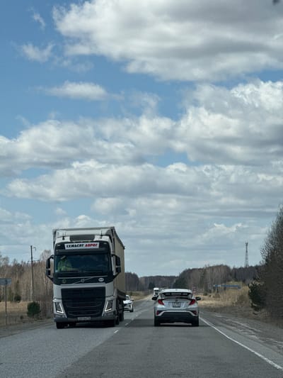 The image shows a road with a white truck and a white car driving on it. The truck has a Russian license plate "K667BA 146". The word "СЕМЬСОТ ДОРОГ" is visible on the front of the truck, which means "Seven Hundred Roads" in Russian. There are trees on both sides of the road. The sky is cloudy with patches of blue. There is a tall tower in the distance. Given the truck's writing "СЕМЬСОТ ДОРОГ" and the license plate, the location is likely to be Russia. However, there is insufficient data to pinpoint the exact location as Mar'evka, Russia.