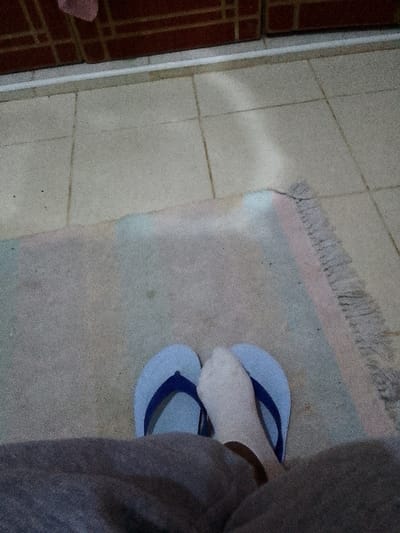 The media file presents a low-angle, top-down view focused on a person's feet and the floor area. The setting appears to be an indoor or covered outdoor space.

The subject is a person, whose lower legs and feet are visible. They are wearing grey, casual trousers, possibly sweatpants. One foot is adorned with a white sock, and both feet are placed loosely inside light blue flip-flops with dark blue straps.

The floor is covered with light-toned, possibly beige or off-white, square tiles separated by grout lines. Partially covering the tiles is a light-colored rug, appearing grey or off-white, with subtle horizontal stripes in pastel shades of light pink/purple and light blue, and a fringed edge on the right side. An elongated patch of bright light, likely sunlight, is cast across the tiled floor and extends onto the rug, suggesting it is daytime.

In the background, a dark reddish-brown structure, possibly a wall with a patterned or brick-like texture, is visible above a narrow white strip that separates it from the tiled floor. There is no specific activity depicted other than the person's feet resting in a relaxed posture. No text is visible in the image. The image has a slightly grainy quality.