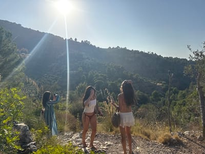 The image depicts three females on a rocky, unpaved trail in a mountainous, heavily vegetated area of Deià, Spain, under bright sunlight. The intense sun, positioned high in the upper-center, produces a prominent lens flare.

On the left, a female wearing a long, blue and white patterned dress is seen from behind, partially obscured by green foliage. She holds a dark handheld device to eye level, appearing to capture an image or video.

In the center, a female in a white tank top and dark red bikini bottoms stands barefoot on the trail, her right hand raised to her face.

On the right, a third female stands barefoot with her back to the viewer, dressed in a white skirt with lace trim and a dark bikini top. Her right hand is positioned at the back of her head, and she carries a woven bag in her left hand.

The immediate foreground features rocks and green shrubbery. The midground and background consist of steep, tree-covered mountain slopes, with scattered distant structures visible on the hillsides. A thin, upright dead branch or stake is visible on the right side of the trail.