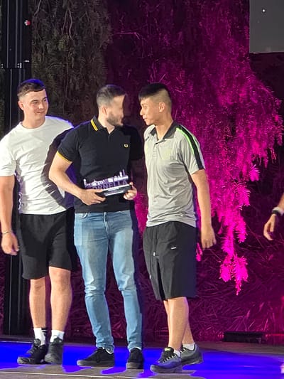 This image depicts an awards ceremony held outdoors, likely in Cornellà de Llobregat, Spain, given the event's branding. Three men are the primary subjects on stage. The man on the left, wearing a white t-shirt and black shorts, is looking to his left. In the center, a man in a black polo shirt with yellow trim and blue jeans is holding an award. He is looking at the man to his right. The man on the right, dressed in a grey polo shirt with green accents and black shorts, is also looking towards the man in the center. The background features lush greenery illuminated with vibrant pink lighting, creating a dramatic ambiance. The stage floor is reflective and illuminated with blue light.