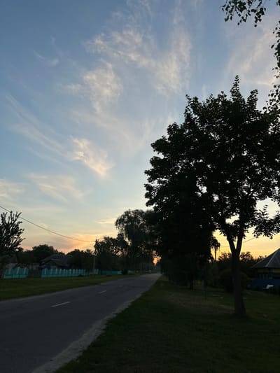 The image depicts a rural road at dusk, with a silhouette of trees on the right side. A power line runs across the frame. In the background, there are houses and a fence. The sky is a gradient of blue and orange, with wispy clouds. The location is identified as Буркоўскі сельскі Савет, Belarus. There are no people visible in the image.