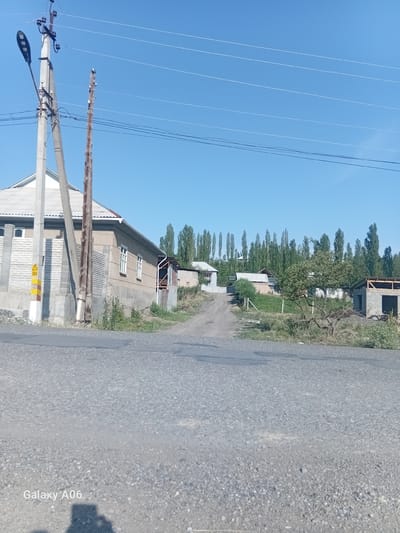 The image depicts a rural scene in the city of Kojo-Aryk, Kyrgyzstan, under a clear blue sky. In the foreground, a grey asphalt road leads into the distance. To the left, a concrete utility pole with a streetlamp and multiple wires is prominently featured. Adjacent to it, a wooden utility pole stands with wires running across the frame.
Further into the scene, a single-story house with a grey roof and white-trimmed windows is visible. Next to this house, a dirt road winds uphill. On the right side of the image, a partially constructed building with cinder block walls and exposed wooden roof beams is present. A mature tree with green foliage is situated in front of this construction.
A dense line of tall, slender trees forms a backdrop across the middle ground. The overall environment suggests a residential area with ongoing development and a natural, verdant landscape. The image is captured during daylight, with the sun casting shadows. A watermark indicating "Galaxy A06" is visible in the bottom left corner.