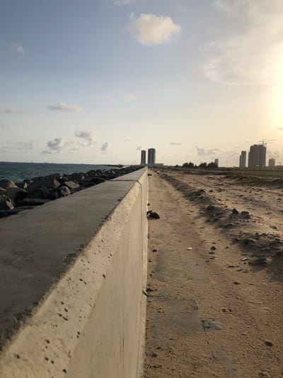 The image depicts a coastal scene in Kuramo Waters, Nigeria. A long concrete barrier runs diagonally across the frame, separating the ocean on the left from a sandy beach on the right. Large rocks are piled along the base of the barrier, acting as a breakwater. In the distance, several tall buildings are visible along the coastline, partially obscured by a hazy sky. The lighting suggests late afternoon or early morning. There are no people visible in the image.