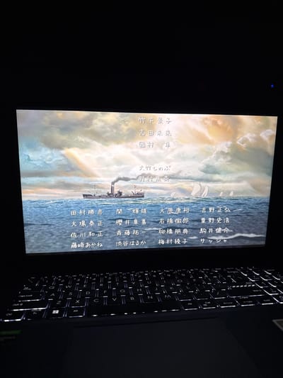 A dark-colored laptop is positioned in a dimly lit indoor environment, viewed from an angled, slightly elevated perspective. The laptop's screen prominently displays a still image depicting a body of water under a cloudy sky with warm, possibly sunset or sunrise, hues. A dark-hulled steamship with a single smokestack emitting smoke is centrally located on the water, with several smaller white-sailed vessels visible in the background. Japanese characters, consistent with movie or show credits, are superimposed over this maritime scene. The keyboard below the screen is illuminated by white backlighting, while the touchpad and palm rest areas remain dark. No individuals are present in the image. The scene is situated in the city of Spitak, Armenia.