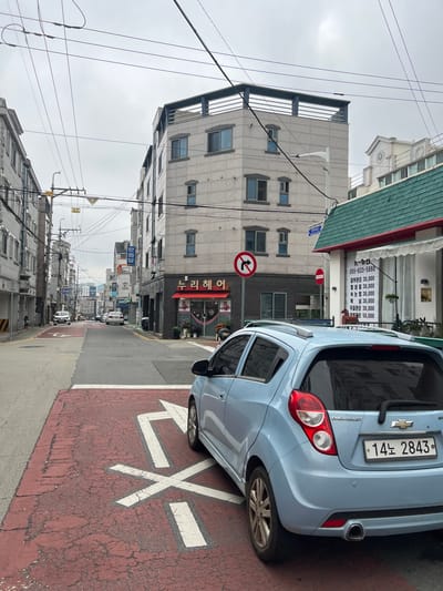 In Geoje-si, South Korea, on an overcast day, a light blue Chevrolet Spark car is parked on a red asphalt road marked with white pedestrian crossing lines. The car, facing away from the viewer, has a license plate reading "14노 2843". Behind the car, a multi-story building with a light-colored facade and dark trim features multiple windows. To the right of this building, a store with a green roof and red awning displays a white sign with Korean text and prices, including "피부관리 30,000" (Skin care 30,000). Across the street, other buildings and cars are visible, along with a dense network of electrical wires overhead. A "no U-turn" sign is posted on a pole on the left side of the street. The scene depicts a typical urban street in a South Korean city.