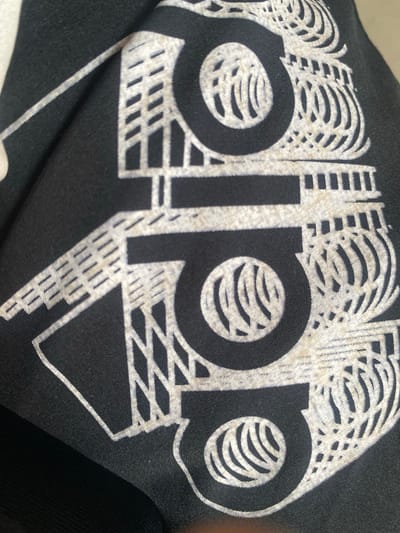The image shows a close-up of a black fabric with a white graphic design. The design appears to be a stylized logo, possibly for a brand. It features bold, curved lines and geometric shapes. Some sections of the design are filled with a pattern of parallel lines, resembling a crosshatch or a grid. The overall impression is abstract and modern. The fabric is creased and folded, indicating it might be clothing or a piece of material. There are no people or a specific scene visible. The lighting suggests an indoor setting or an overcast day, as there are no strong shadows. The color palette is limited to black and white with some muted, off-white tones within the graphic itself, possibly due to the printing process or age of the fabric. No text is clearly discernible within the graphic.