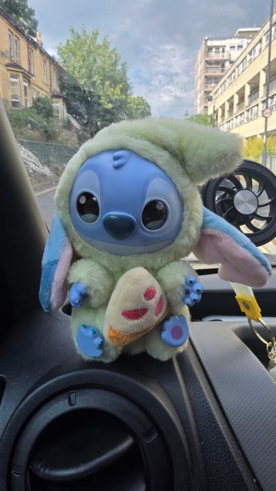 A plush toy of the character Stitch, dressed in a fuzzy pale yellow hooded outfit with pastel blue and pink bunny ears, is positioned inside a vehicle. The toy has large, expressive eyes and is a light blue color. It is lying on its back, with its limbs outstretched. Attached to the toy is a gold keychain with a yellow strap.
The setting appears to be the interior of a car, with textured dark grey and black dashboard elements visible. The toy is placed near an air vent. Through the car window, a view of buildings and trees can be seen, suggesting an urban or suburban environment. The reflection on the window includes details of buildings with multiple windows and a sign with red lettering, possibly a road sign. The sky is cloudy, indicating overcast weather. The overall impression is a casual snapshot of a car's interior with a decorative item.
The image is taken from a slightly elevated angle, looking down at the toy. The lighting suggests it is daytime, but the overcast sky prevents strong shadows. The orientation of the image is such that the toy is upright and the background is horizontal, implying the original photo was taken with the camera rotated. There is no visible text on the plush toy itself, but a red and white sign is visible in the reflection on the window. The context provided states the location is Bristol, United Kingdom.