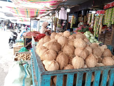This is an outdoor market scene in the city of Uruekanoe, Indonesia, featuring a stall selling various fresh produce. In the foreground, a large blue wooden crate is filled with a bounty of coconuts. Behind the coconuts, a person wearing a light-colored hijab is visible, tending to a display of red chilies and what appear to be shallots. Further back, to the left, a motorcycle is parked. The stall is covered by a colorful striped awning, and hanging above are various bags and packets of goods, likely spices or seasonings. To the right, a large stack of egg cartons is visible. The ground surface appears to be dirt or gravel. The overall impression is a vibrant, bustling local market.