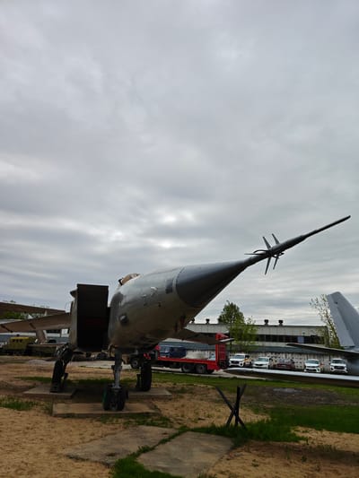 A low-angle, daytime shot depicts an exposed Tupolev Tu-22M3 bomber at the Vadim Zadorozhny Technical Museum in Arkhangel'skoye, Russia. The aircraft, primarily painted gray, is resting on concrete slabs amid a sandy ground interspersed with patches of green vegetation. Its landing gear is deployed. Behind the bomber are assorted vehicles, including a red flatbed truck and several cars parked in a row. In the background, a low building with a flat roof and evenly spaced windows is visible. The sky is overcast.