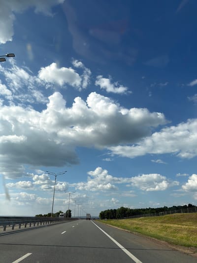 The image depicts a highway in the city of Shali, Russia, under a bright blue sky filled with fluffy white clouds. The perspective is from a vehicle moving along the road, looking forward. The asphalt road has clear white lane markings. On the left side of the road, there is a metal guardrail, and beyond that, a body of water or a wide expanse. Several lampposts line the left side of the highway. On the right side of the road, a grassy embankment slopes upwards, topped by a fence and a dense line of green trees. In the distance, a white truck is visible on the highway, and another vehicle can be seen further ahead. The scene is one of daytime travel on a sunny day.