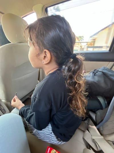 A young girl with dark brown, curly hair tied in a ponytail is sitting in the back seat of a car. She is wearing a black long-sleeved shirt and striped pants. Her left hand is holding a thin, brown stick-like object. The car's interior is visible, with beige leather seats and a window showing a blurry outdoor scene. There is a black bag behind her. The location is the city of Kedungreja, Indonesia.