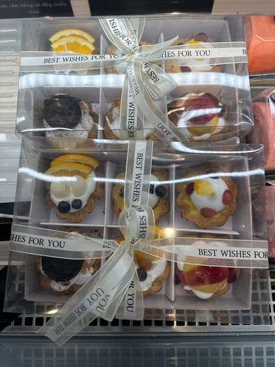 The image displays two stacked clear plastic boxes, each containing six small pastries. The pastries are arranged in a 2x3 grid within each box. Each box is secured with a white ribbon printed with the words "BEST WISHES FOR YOU" in black lettering. The pastries themselves vary in decoration. Some are topped with white cream and fruit slices, such as oranges and what appear to be berries, while others have an Oreo cookie or a chocolate topping. The overall presentation suggests a bakery or gift shop setting, possibly in Phu Thuy, Vietnam, given the text in the background which appears to be Vietnamese. The lighting is bright, highlighting the details of the pastries and packaging.