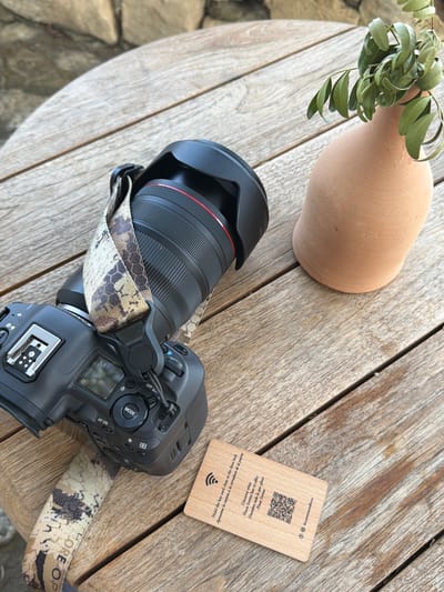 An overhead shot captures a wooden table with a camera and a terracotta vase containing green leaves. The camera, a black DSLR with a camouflage strap, rests on the left side of the table. The strap has the words "EXPLORE" and "OP" visible. A small wooden card lies in front of the camera. The card features text and a QR code, with a Wi-Fi symbol and Instagram logo. The terracotta vase, a light brown color, sits on the right side of the table, holding a small cluster of green leaves. The table surface shows the texture of weathered wood planks. In the background, out-of-focus stones are visible, suggesting an outdoor setting. The overall impression is one of a still life arrangement, possibly in Montuïri, Spain, given the context of the card's text referencing "Finca Serena".
