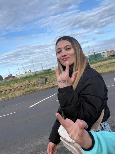 An outdoor, eye-level, daylight photograph captures two individuals against an industrial backdrop. A young woman with long, straight, light-brown hair and light makeup occupies the mid-ground, facing towards the viewer and slightly to the left. She wears a black puffer jacket and light-wash blue jeans, with a beige "GUESS" branded strap visible across her chest. Her left hand is raised, forming a "rock on" gesture with the thumb, index, and pinky fingers extended, while the middle and ring fingers are curled. The side of her left palm bears a black tattoo reading "Tavia" (Távia) in Greek script. Her lips are slightly pursed, and she has a subtle smile.

In the immediate foreground, partially visible on the bottom right, is the outstretched right arm and hand of a second individual, wearing a light-blue long-sleeved garment. This hand has two fingers extended towards the camera in an unclear gesture.

The background features a grey asphalt road with a solid white line and a broken white line. Beyond the road is a grassy, unkempt embankment with dry vegetation. A rusted metal railing runs along the base of the embankment, and a weathered concrete structure is partially embedded in the grass. In the further background, under a partly cloudy sky with patches of blue, an expansive industrial complex is visible. This complex includes multiple low-lying buildings with light-colored roofs, several tall poles or masts, and various cranes, including one red gantry crane and numerous other industrial lifting structures, strongly indicative of a port or shipyard. Two large, dark-green cylindrical storage tanks are also discernible on the right side of the background, along with other industrial buildings featuring chimneys.

Based on the industrial characteristics, particularly the presence of port cranes and associated structures, the scene is situated in an industrial area, most likely around the Port of Boston, United Kingdom.