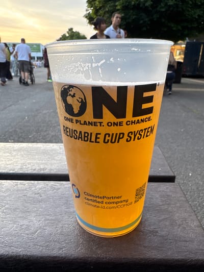 A close-up, slightly tilted, medium-shot photograph shows a filled, reusable yellow cup with black lettering that reads "ONE. ONE PLANET. ONE CHANCE. REUSABLE CUP SYSTEM." The cup has a white rim and a green band at the bottom. The text "ClimatePartner certified company climate-id.com/CCFIG8" is printed near the bottom of the cup. A QR code is also visible on the cup. The cup sits on a dark gray, textured surface. In the blurred background, people stand and walk in an outdoor area. Trees and buildings are visible in the distance, with an early evening or late afternoon sky suggesting dusk or dawn. Given the certification from ClimatePartner and the outdoor setting, this photo likely represents an environmental initiative at a local Borough of Spelthorne event in the United Kingdom.