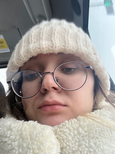 A close-up, front-facing self-portrait depicts an individual, likely female, situated inside a vehicle. The subject wears a white, ribbed knit beanie that covers the forehead and ears. Large, round, metallic-framed glasses obscure the upper face, revealing green eyes. A small, dark nasal piercing is visible on the left nostril. The lips are closed, exhibiting a neutral expression. A white, textured, fleecy material, consistent with a collar or scarf, surrounds the lower face and neck. Portions of light brown or blonde hair are visible beneath the beanie. The background indicates an interior space, with a light-colored overhead panel and a partially visible yellow label in the upper left. The upper right shows a blurred exterior through a window, including a dark vertical support and a faint green light, possibly a traffic signal. The precise location within the city of Борисово, Russia, is not discernible from the visual evidence.