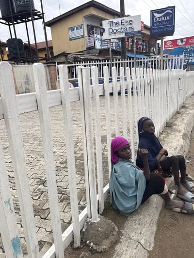 The image shows two young people resting on a white metal fence in what appears to be a busy urban area in Lagos, Nigeria. The person on the left is wearing a dark blue hijab and a dark blue top, with their arms crossed. The person on the right is wearing a light blue garment and a pink hijab, with their head resting on their arms. Their expressions are calm, suggesting a moment of respite.
The background reveals a commercial street with various signs and buildings. Prominent among the signs are "Dulux let's colour colour shop" and "The Eye Doctors Eye Care We Care". Other visible text includes "PRINT ARENA" and "itel Energy", along with some signs in what appears to be Nigerian languages. The setting is cluttered, with visible electrical cables, water tanks, and other urban infrastructure. The ground is a mix of concrete and dirt. The overall atmosphere is that of a typical day in a bustling Nigerian city, with the individuals finding a quiet spot to rest amidst the activity. The lighting suggests it could be daytime, possibly overcast given the light.