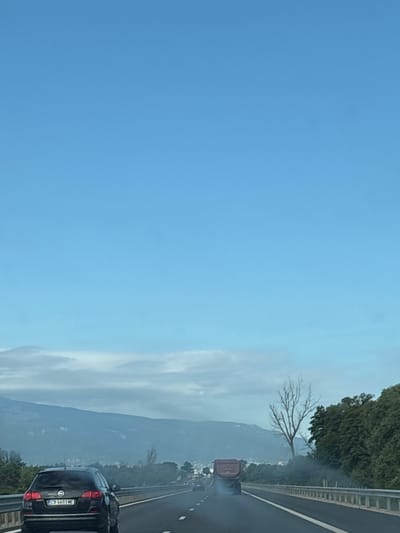 The image depicts a highway scene with a clear blue sky. In the foreground, a black station wagon is visible on the left lane of the road, with its license plate reading "CB 6603 ME." Further ahead on the right lane, a red truck is emitting a trail of smoke. The road is bordered by a metal guardrail and lush green trees on the right. In the background, a mountain range is shrouded in a light mist or fog, and a bare tree stands prominently on the right side of the road. The overall impression is that of a journey through a semi-urban or rural landscape. The scene appears to be located on the outskirts of Dolni Bogrov, Bulgaria.