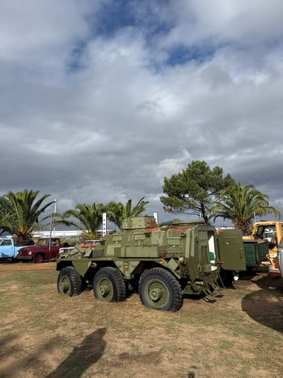 The image shows an outdoor scene in Kraaifontein, South Africa. Dominating the view is a line of vintage military vehicles parked on a grassy field. In the foreground, the prominent vehicle is a large, olive-green armored personnel carrier with six wheels. Behind it are several older civilian vehicles, including a red pickup truck and a blue truck. Scattered palm trees are interspersed amongst the vehicles and along the background. The sky above is overcast with dark storm clouds.