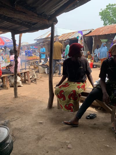 The image depicts an outdoor market scene in Dull A, Nigeria. In the foreground, two individuals are seated on a wooden bench. The person on the left wears a dark, graphic t-shirt and a vibrantly patterned skirt with red and green motifs. They are looking to the right. The person on the right is partially visible, wearing dark trousers and a patterned garment. Their legs are crossed, and they are wearing flip-flops.

Behind them, a bustling marketplace unfolds. Several stalls with corrugated tin roofs are visible, some appearing weathered. A variety of goods are displayed, including packaged items in blue and yellow boxes, and bottles on shelves. People dressed in traditional clothing are milling about. One person in a light blue garment is seen in the background, and another in a light green garment with a cap. There are also signs visible, including one with red and blue branding. The ground is predominantly dirt, with some scattered debris and wooden structures. The overall atmosphere suggests a lively, everyday market environment.