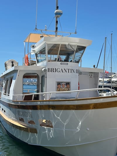The image shows the boat "Brigantin II" docked in the marina of Saint-Tropez, France. The boat is white with a wooden trim along the deck. The name "Brigantin II" is written in large black letters on the side of the boat. On the side of the boat, there are three framed images of people in uniform, appearing to be mannequins or painted figures. The sky is clear and blue, and other boats are visible in the background.