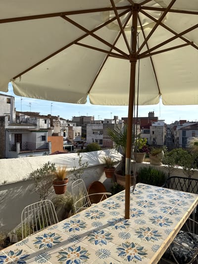 This media file showcases an outdoor terrace scene overlooking a dense urban landscape. The foreground is dominated by the underside of a large, light-colored patio umbrella, its wooden spokes and central pole clearly visible. To the right, a table with a decorative tiled surface featuring a repeating floral pattern in blue and white is prominently featured. The table is set against a backdrop of various potted plants and white metal chairs.
The mid-ground offers a view of a collection of buildings, likely apartments or houses, packed closely together. The architecture appears to be typical of a Mediterranean town, with a mix of white, terracotta, and grey facades, some with visible brickwork. Rooftop antennas are common, suggesting a residential area.
The scene is bathed in bright sunlight, indicating it is daytime, likely late morning or early afternoon given the quality of the light. The sky is clear with a few wispy clouds. The overall impression is one of a relaxed, sunny day in a European coastal town. There are no people or explicit activities depicted, but the presence of the umbrella, table, and chairs suggests a setting intended for leisure and outdoor dining or relaxation. The location context provided, Vilanova i la Geltrú, Spain, further supports the visual cues of a Mediterranean setting. No visible text is present in the image.