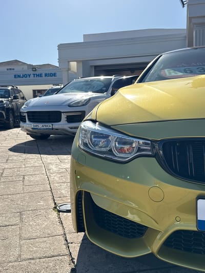 The image displays a close-up, low-angle view of the front of a vibrant yellow-gold sports car, likely a BMW M4, parked outdoors on a bright, sunny day. Its detailed headlight, aggressive black grille inserts, and front bumper with circular parking sensors are prominent in the foreground.
Behind this car, a white Porsche Macan SUV is parked, also facing forward, revealing its grille and a number plate frame that reads "CARS4AFRICA". A black vehicle is partially visible further behind the Porsche. The cars are situated on a grey, block-paved surface.
The setting appears to be a car dealership or a similar commercial establishment. In the background, a white building with visible architectural details displays the blue text "ENJOY THE RIDE" on its facade. Strong shadows are cast on the ground, indicative of direct sunlight, suggesting a clear day during midday or early afternoon.
Visible text includes "ABS 63" and "APPLY" on a sticker on the yellow car's windshield, "CARS4AFRICA" on the white car's number plate frame, and "ENJOY THE RIDE" on the building. A faint "R200 995" is also visible on the white car's windshield, likely a price tag. No people are present in the scene. The context suggests a location in Blouberg, South Africa.