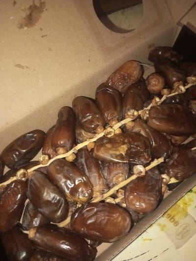 The image depicts an open, light-brown cardboard box containing a large quantity of dark brown dates. The dates are mostly oval-shaped, visibly wrinkled, and possess a slight sheen. Many individual dates remain attached to short, light-colored stem segments and dried calyxes. The dates are piled densely, occupying the majority of the box's interior. The cardboard box exhibits visible folds and a circular cut-out in its upper right quadrant, potentially for handling or ventilation. In the upper left quadrant of the interior cardboard surface, several small, irregular dark stains are present. A white label with partially visible English text, including "DATES" and "HORCHATA," is visible beneath some dates in the bottom right corner of the box. No individuals are present in the scene, and no actions or interactions are depicted. The scene is located in the city of Ura-Dozhi, Bhutan.