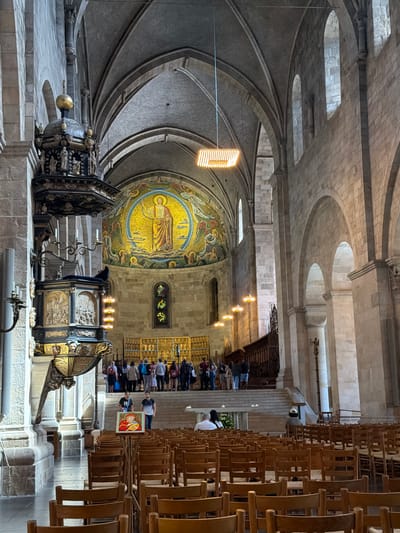The image shows the interior of Lund Cathedral in Lund, Sweden. The cathedral features a large, circular mosaic above the altar depicting Christ. In the foreground, rows of wooden chairs fill the nave. A pulpit, ornately carved and decorated, stands to the left of the frame. Several people are visible throughout the cathedral, some standing in groups, others seated in the chairs, and a few walking. Natural light streams in through arched windows high on the walls, illuminating the stone architecture and the golden mosaic. The overall impression is one of historical grandeur and serene atmosphere.