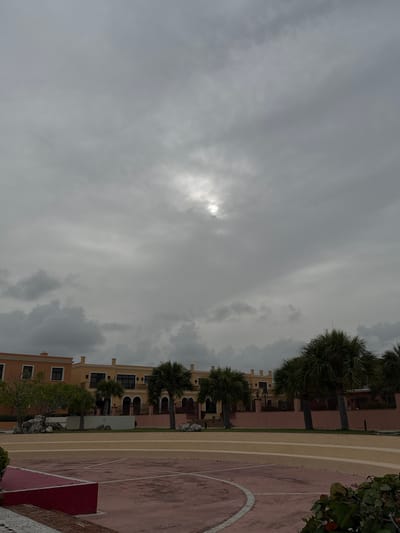 This image depicts a cloudy sky over a residential area in Higüey, Dominican Republic. The sky is filled with dense gray clouds, with a faint sunlight visible through a break in the clouds on the left side of the image. A line of palm trees runs along the right side of the frame, separating the sky from the buildings and a sports court.
The buildings are multi-story structures with light-colored facades, featuring balconies and arched windows. The sports court, possibly a basketball court, is partially visible on the far right, with its red and pink surface marked with white lines. The overall scene suggests a possibly humid or overcast day. No people are visible in the frame, and there is no visible text. The composition, with the sky dominating the left half and the buildings and court on the right, creates a sense of depth and place. The presence of palm trees and the architectural style of the buildings are consistent with a tropical location like the Dominican Republic.