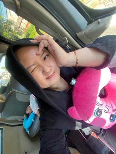In Gayaman, Indonesia, a woman in a black hijab is taking a selfie in a vehicle. She is wearing a gold bracelet and has her hand on her forehead. Next to her, a pink stuffed animal with large blue eyes and eyelashes rests on her shoulder. In the background, a child is sleeping in a car seat, adorned with a blue neck pillow. The scene appears to be during daylight, with sunlight streaming through the vehicle's windows.