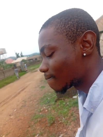A close-up, side-profile photograph shows a young Black man with short, dark hair and a light beard. He is wearing a light blue collared shirt, and a small, silver stud earring is visible in his left ear. His eyes are closed, and he appears to be looking down and slightly away from the camera. The man's face is the primary subject of the image.
The background is a blurred outdoor scene. A dirt road with tire tracks runs diagonally across the frame from the bottom left to the middle right. Green grass and patches of dirt are visible on either side of the road. In the distance, there are some low-lying buildings, a water tower on a pole, and lush green hills under an overcast sky. The overall impression is of a rural or semi-urban environment. Based on information from the user, the location is identified as Ashata, Nigeria. No specific actions or interactions are clearly depicted, as the focus is on the portrait of the man.