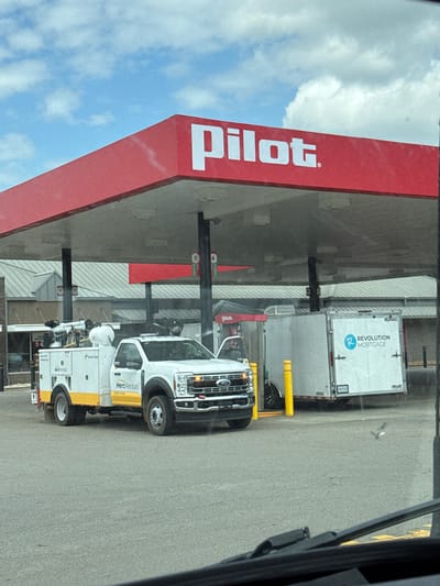 The image shows a Pilot gas station with a red canopy. Under the canopy, a white service truck with "HercRentals" written on it is parked, along with a white trailer. The trailer has "Revolution Mortgage" logo on it. The ground is asphalt, and the sky is cloudy blue. The image appears to have been taken from inside a vehicle, as evidenced by the windshield wipers and dashboard in the foreground. There are no people visible in the image. Based on the Pilot branding and typical locations for such businesses, this scene is likely in Mebane, United States of America.