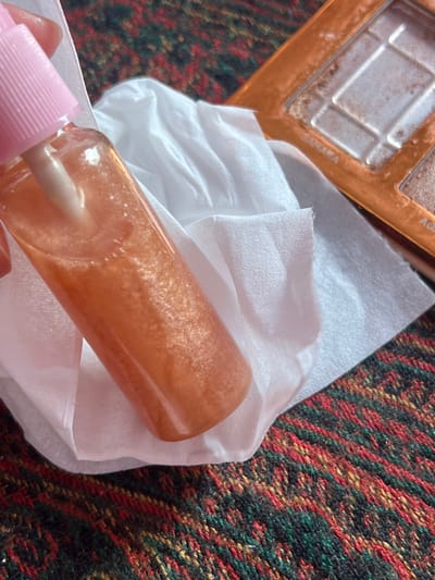 This image, captured in Ksar-el-Kebir, Morocco, presents a close-up, downward-angled view of several cosmetic items resting on a patterned textile surface. A clear plastic spray bottle containing a shimmering, orange-gold liquid with visible particles is held vertically in the left foreground by human fingers. The bottle features a light pink pump top and nozzle. Directly beneath the bottle and extending to the right is a crumpled white tissue. To the upper right, an open makeup palette with an orange-gold-colored case is partially visible. This palette contains multiple compartments of light-toned, shimmering powder, some labeled "SAHARA" and "AGA," showing clear signs of prior use. The background consists of a tightly woven textile, likely a blanket or rug, displaying a pattern of dark green, red, yellow, and black threads.