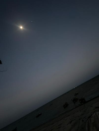 The image displays a tranquil nighttime scene, likely along a coastal or riverside area in Bagamoyo, Tanzania. The dominant feature is a bright, luminous moon positioned in the lower-left portion of the dark sky. To the upper-left of the moon, a distinct, bright star or planet is visible. The sky transitions from a deep indigo-black at the top and left to a lighter, hazy grey across the horizontal middle of the frame, suggesting atmospheric mist or distant light pollution near the horizon.
On the far right side of the image, a dark, textured wall or rocky bank extends vertically, appearing silhouetted against the hazy backdrop. Several indistinct, darker shapes are discernible on or near this structure, possibly representing small boats, debris, or other objects. A faint, skeletal tree branch is barely visible in the very bottom-left corner, silhouetted against the moonlit sky.
The scene depicts nighttime conditions with a clear upper sky contrasting with a misty or foggy layer near the horizon. No explicit activity is taking place, and there is no visible text.