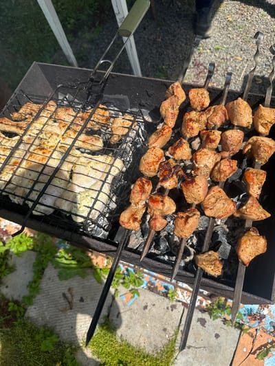 In Мазалаўскі сельскі Савет, Belarus, an outdoor cooking scene unfolds. A black metal grill is laden with food. On a wire rack on the left, several pieces of seasoned chicken are being cooked, appearing pale and lightly browned. To the right, on metal skewers, chunks of seasoned pork are grilling over hot coals. The pork pieces are a golden brown, some showing slight charring. The grill is positioned on a surface with patches of green vegetation and concrete. The overall impression is a rustic, sunlit barbecue in progress.