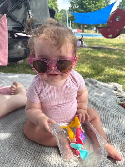 A baby is lying on a grey blanket outdoors, possibly at a park or playground in Giessen, Germany. The baby is wearing a pink ribbed onesie and pink glittery sunglasses. The baby is holding a clear plastic container with small mermaid-like toys inside. In the background, there is green grass, trees, a blue sail shade, and a red circular object. A stroller is partially visible to the left of the baby, with the word "Joie" clearly visible on it. The scene appears to be set during the daytime, with bright sunlight casting shadows, suggesting fair weather. The baby's gaze is directed towards the viewer, and their expression seems curious or playful. The image is taken from a high angle, looking down at the baby.