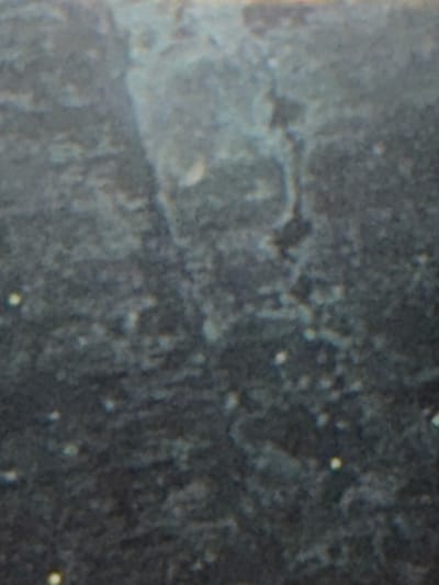 The image shows a close-up, out-of-focus view of a dark, textured surface, likely a wall or ground, with several small, bright white dots scattered across it. There is a lighter, vertical, irregular shape in the upper center of the image, possibly a crack or a natural feature in the material. The overall impression is one of grit and imperfection. The specific location in Wildwood, United States, is not identifiable from the visual information provided.
